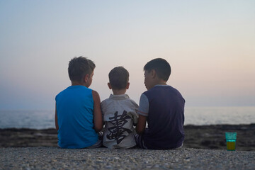 Three boys sitting on the shore of the sunset sea, rear view