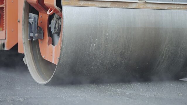 Steamroller compactor cylinder flatten out the new asphalt with steam evaporationg in slow motion. Industrial construction concept