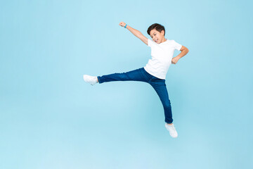 Cute mixed race Asian kid boy playing by jumping with wide open legs and raising fist in light blue...