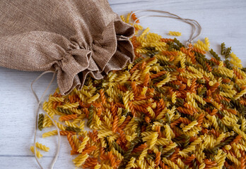 Uncooked multicolored Fusilli pasta spilled out of fabric bag over white wood background. top view.