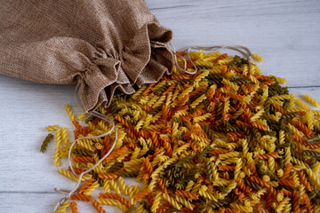 Uncooked multicolored Fusilli pasta spilled out of fabric bag over white wood background. top view.