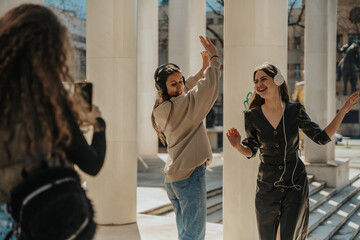 Curly haired girl taking a photo of her two brunette friends listening to music on their headphones, dancing and singing