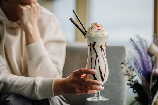 Close up image of a woman's hand holding a glass of ice cream cocktail with cream on top, light bokeh 