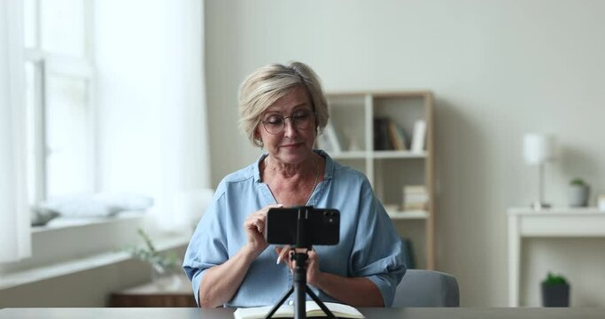 Cheerful senior influencer, blogger, coach woman recording selfie video, sitting at table, speaking at smartphone fixed on tripod, smiling, broadcasting online, giving workshop to blog audience