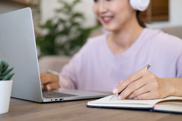 Close up hand - Attractive female student learning language, watching online webinar, listening audio course, Focused woman wearing headphones using laptop writing notes, e-learning education concept