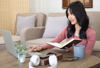 Young woman in casual at wooden table in living room and holding notebook . In front of her is laptop, smartphone. Student learning online with computer doing research or preparing for exam online.