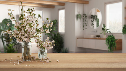 Fototapeta na wymiar Wooden table, desk or shelf close up with branches of cherry blossoms in glass vase over blurred view of modern bathroom with houseplants, urban jungle interior design concept