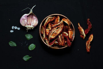Dried tomatoes in wooden bowl and levitating tomatoes above, on the black background with garlic and basil leaves