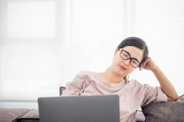 Asian mature woman wearing glasses seriously working on computer laptop sitting on sofa couch in living room house. She thinking find solution problem of work.