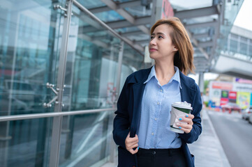 Beautiful Woman Going To Work With Coffee Walking Near Office Building. Portrait Of Successful Business Woman Holding Cup Of Hot Drink In Hand On Her Way To Work On City Street. High Resolution.