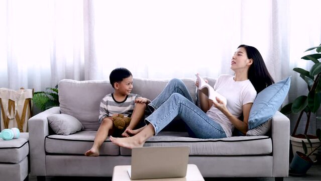 Asian beautiful mother and son read favorite book together on weekends, young woman and adorable boy lying down on sofa reading a comic book in the living room with happiness and fun.