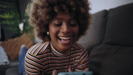 Video of smiling African American pretty woman using cell phone lying on sofa in living room. Happy young black afro girl flirting on mobile dating app at home. People addicted social media.