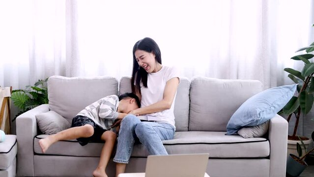 Happy Asian mother stop read a book and inviting adorable son tickle play tickle in living room. Nanny play tickle with little preschool kid laughing having fun lying on sofa, enjoy game togethe