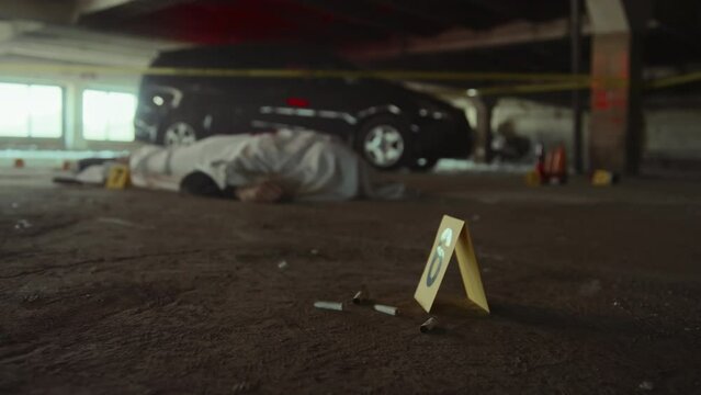 Full selective focus shot of murder scene, body of victim under white sheet, red and blue police lights reflecting on walls and parked car, cigarette butts and forensic marker in foreground
