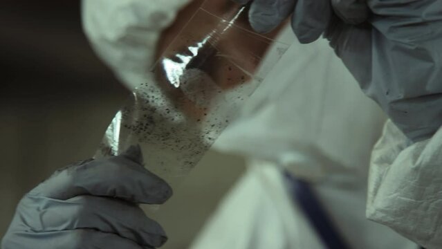 Close-up shot of face and hands of female forensic investigator in hooded overalls and mask carefully examining fingerprints in dust on piece of transparent adhesive tape, while investigating murder