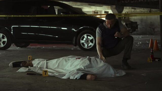 Full shot of Caucasian male police detective, in bulletproof vest and holding torch, investigating murder scene at dark underground parking lot and inspecting dead body covered with white sheet