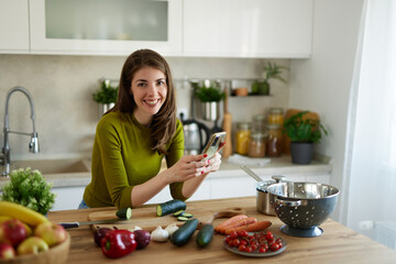 Young woman text messaging while cutting vegetables on cutting board in the kitchen