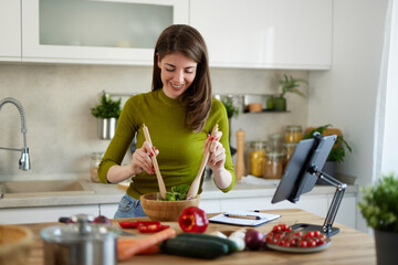 Young woman standing in the kitchen making a salad using online recipe