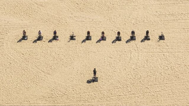 Aerial 4K shot of people cycling on bicycle trainer. Bicycle sports simulators lined up on the sandy beach with trainer showing exercise and motivating people for healthy life wellbeing, Santa Monica
