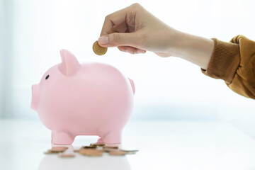 Woman hand putting money coin into pink piggy bank on white table for saving money wealth and financial concept.