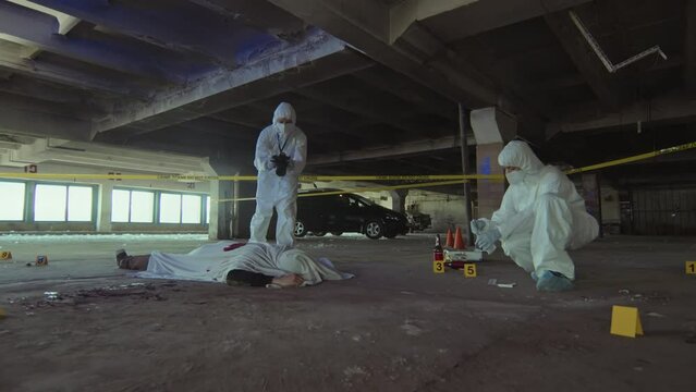 Full slowmo shot of two unrecognizable forensic investigators working on murder case in abandoned garage, one taking photographs of corpse on camera, and colleague collecting samples and evidence