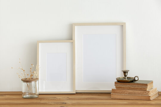 Horizontal wooden frame mockup on a vintage wooden table. Vintage candlestick on books, vase with white dry grass and flowers on white wall background. Scandinavian room interior design. 
