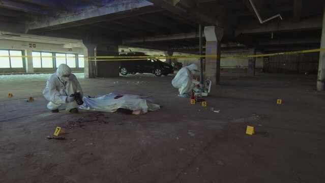 Full arc shot of unrecognizable male or female forensic specialist in white suit and mask crouching on ground and taking photographs of crime scene beside corpse, and colleague collecting samples