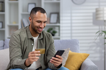 Smiling young African American man sitting on sofa at home and using credit card and phone, checking account, making money transfer, online shopping