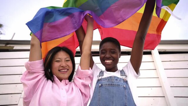 Chinese and African women raising a rainbow flag outside during day, looking camera happily in honor of the LGBT pride month. Lesbian multiethnic couple celebrating.