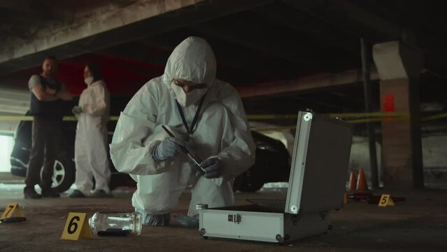 Full shot of unrecognizable forensic expert in protective suit, mask and gloves, dusting empty bottle with brush and powder looking for fingerprints and evidence, and colleagues talking in background
