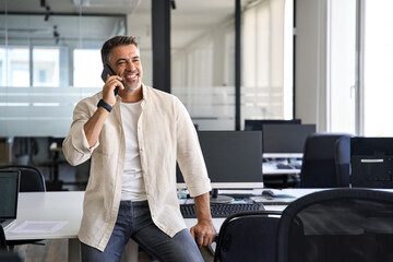 Middle aged Latin or Indian businessman having call on smartphone with business partners or clients. Smiling mature Hispanic man in relaxing pose talking by mobile cellphone at work in modern office. 