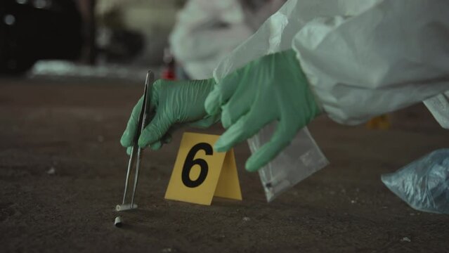 Close-up handheld shot of hands of unrecognizable forensic expert in protective suit and gloves gathering cigarette butts from ground using forceps, putting into ziplock bag, sealing it and leaving