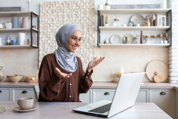 Young smiling Muslim woman in hijab sitting in kitchen at home and talking on video call via laptop.