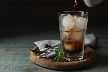 Pouring coffee into glass with ice cubes, mint and beans on light blue wooden table, space for text