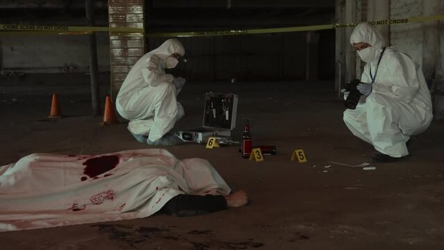 Full shot of body of murder victim on ground under white sheet with blood stains, and two unrecognizable male or female forensic experts taking photos of crime scene and collecting evidence