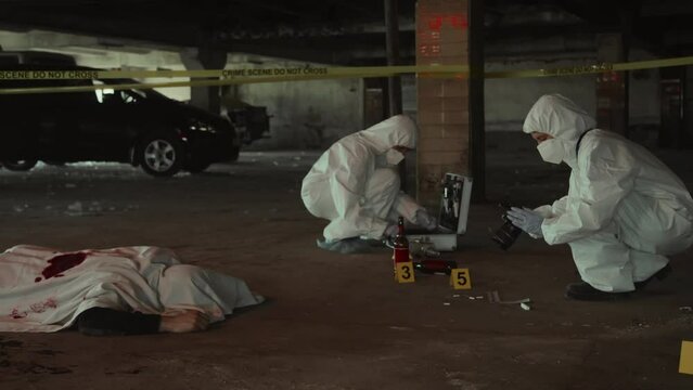 Full shot of two unrecognizable male or female forensic experts in white overalls working together on murder case near corpse, one collecting samples and another taking photographs of crime scene