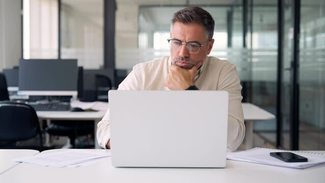 Mature Indian or Latin business man ceo trader using computer, typing, working in modern office, doing online data market analysis, thinking planning tech strategy looking at laptop with copy space.