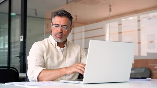 Mature Indian or Latin business man ceo trader using computer, typing, working in modern office, doing online data market analysis, thinking planning tech strategy looking at laptop with copy space.
