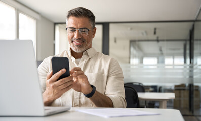 Middle aged Hispanic business manager ceo using cell phone mobile app, laptop. Smiling Latin or Indian mature man businessman holding smartphone sit in office working online on gadget with copy space. - Powered by Adobe