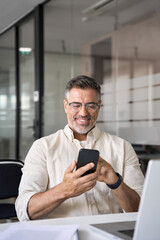 Vertical portrait of middle aged Hispanic business manager using cell phone mobile app. Smiling Latin or Indian mature man businessman holding smartphone sit in office working online on gadget.