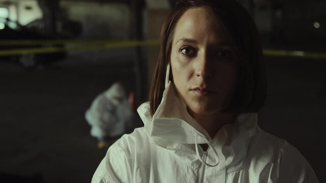 Close-up portrait shot of grim Caucasian female forensic specialist in white overalls and mask hanging off ear posing in front of crime scene and looking at camera, and colleague working in background