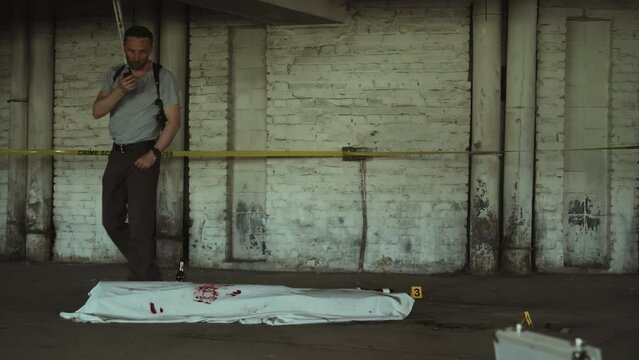Full shot of Caucasian male detective walking around corpse of murder victim lying on floor in underground garage, covered with white sheet with blood stains, and talking into portable radio