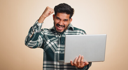 Excited happ young indian man typing on laptop computer, working on project, enjoying results, winning lottery, celebrating success online shopping, win. Hindu guy isolated on beige studio background