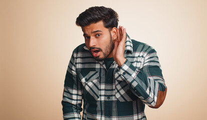 Fototapeta premium I cant hear you. What. Young indian man trying hear, looking confused, frowning, keeping arm near ear for louder voice, asking to repeat, to hear information, deafness. Hindu guy on beige background