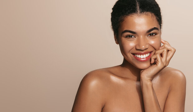 Happy young African American woman with bare shoulders laughing against brown background while representing cosmetology and skincare industry