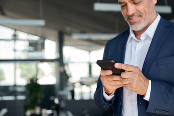 Smiling mid aged business man wearing suit standing in office using cell phone. Mature businessman...