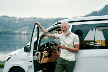 Happy older man standing near rv camper van on vacation using mobile phone. Smiling mature active...