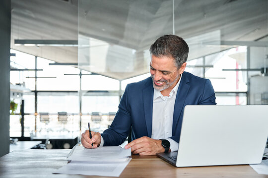 Happy mid aged professional business man executive ceo or lawyer wearing suit sitting at desk signing legal contract document putting written signature authorizing corporate deal in modern office.