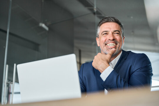 Happy middle aged professional business man company executive ceo manager wearing suit sitting at desk in office working on laptop computer laughing at workplace. Authentic candid photo.