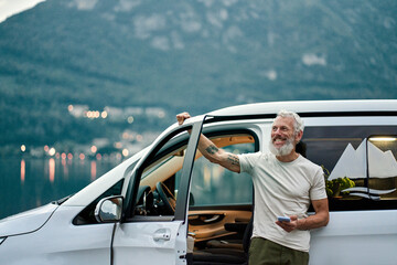 Happy older man standing near rv camper van on vacation using mobile phone. Smiling mature active traveler holding smartphone enjoying free roaming in camping tourism in nature park feeling freedom.